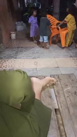 The video shows a person's bare foot and leg in the foreground, wearing green pants, resting on a wooden plank. In the background, four children and two adults are visible. One child, wearing a purple dress, stands to the left. Two children, one in a grey and blue outfit and the other in a yellow outfit, are near an orange plastic chair. An adult, wearing a grey shirt and blue shorts, stands to the far right. Another adult, wearing a yellow outfit, is next to the child in yellow. The environment appears to be an outdoor, possibly residential area, with a tiled floor and some potted plants visible. The lighting suggests it is evening or nighttime. The children seem to be interacting with each other and the chair.