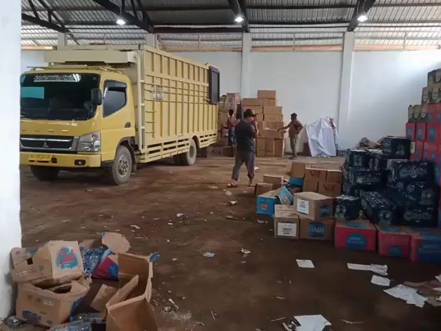 A low-angle, panning shot inside a warehouse-like building shows several people loading or unloading a yellow truck. The interior is large with a metal roof and numerous white pillars. The floor is partially dirt and littered with discarded cardboard boxes and trash. Stacks of packaged goods are visible throughout the space. The overall atmosphere suggests a busy, somewhat cluttered storage or distribution area.
