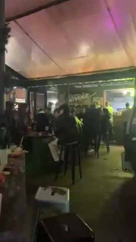 The video begins in what appears to be an outdoor seating area of a bar or restaurant. People are seated at tables and standing, generally socializing. The camera moves to the left to show the interior of a covered outdoor space with exposed brick walls. A person wearing a red robe and a yellow balloon on their head is seen standing in front of a dartboard on the brick wall, throwing darts. Other people are milling around in the background. The atmosphere seems festive and casual.