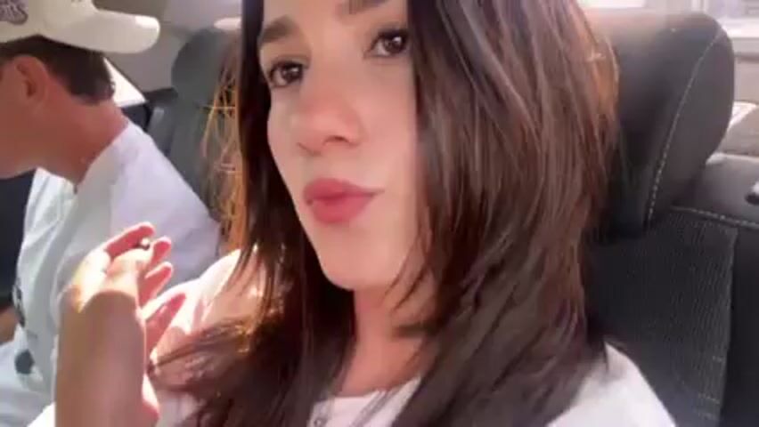 A woman with brown hair sits in the passenger seat of a car, looking directly at the camera. A man in a white shirt and cap is partially visible in the driver's seat. The environment appears to be sunny and inside a vehicle. The woman raises her hand slightly.