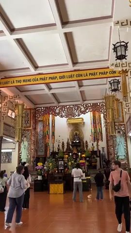 The video shows the interior of a temple. The camera is positioned at a medium distance, facing the altar. The altar is decorated with flowers, candles, and statues of Buddhist figures. In the center of the altar is a large statue of Buddha. Above the altar, a banner with Vietnamese text is displayed. Several people are present in the temple, standing or walking around. They appear to be visitors or worshippers. The lighting is bright, and the overall atmosphere seems peaceful.