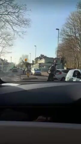 From the interior of a car, a view of a city street in daylight. The camera looks forward towards a traffic light, an intersection, and a building on the right. Cars are lined up on the street, waiting for the light to change. A person on a bicycle is walking in front of the car. The car moves forward as the video progresses, driving under a brick archway. There are trees on either side of the road.