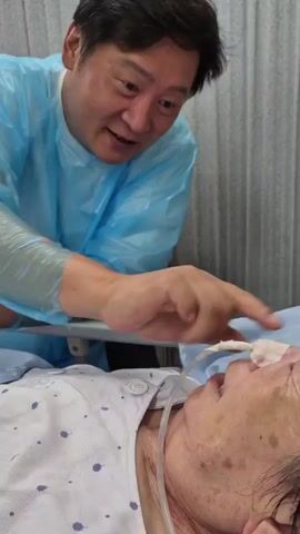 A man wearing a light blue disposable medical gown and what appears to be a protective sleeve on his left arm is leaning over a patient lying in a bed. The patient, whose face is partially visible, has a feeding tube inserted into their nose. The man is speaking to the patient and gesturing with his right hand, which is in focus and held close to the patient's face. The background consists of a light-colored curtain or wall. The overall environment appears to be a hospital room.