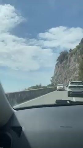 The video shows a view from inside a car driving along a road next to a high, rocky cliff on a sunny day. The sky is blue with scattered white clouds. There are several cars and a motorcycle visible on the road ahead, traveling in the same direction. A stone wall runs along the edge of the road nearest the viewer. The car's dashboard is visible at the bottom of the frame.