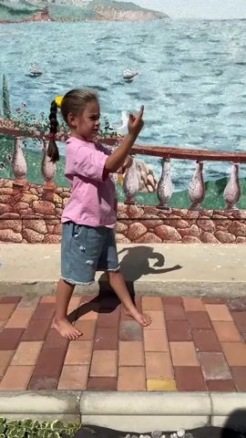 A young girl with a ponytail and a yellow hair tie stands barefoot on a brick pathway. She is wearing a pink t-shirt and blue denim shorts with ripped details. The girl is facing to her right, with her left arm extended and her finger pointing forward. Behind her is a mural depicting a seascape with a sky, water, boats, and a railing with ornate posts. The mural is set against a wall. The girl's shadow is visible on the ground in front of her.