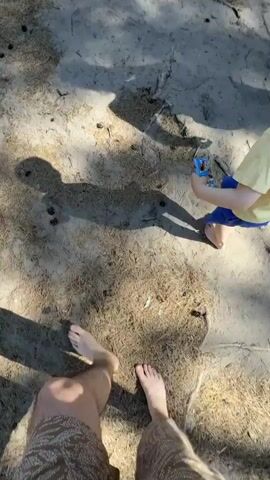 A person is filming themselves and a young child walking barefoot along a sandy path. The path leads through a forest on a sunny day, with trees lining both sides and a fence visible. The child is wearing a yellow shirt and blue shorts and carries a small toy. The person filming is wearing patterned shorts. The camera's perspective is looking down at the ground and then up along the path.