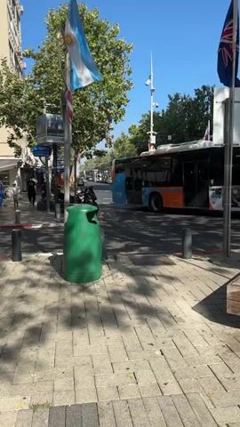 A sidewalk scene in daylight features a green trash can in the foreground. Two flags, one light blue and white, and another with stars and a Union Jack, are visible on flagpoles. A bus with orange and blue sections is parked on the street, with its doors open. Several people are walking on the sidewalk, and trees line the street. Another flag, red and green, is visible on the left side of the frame.