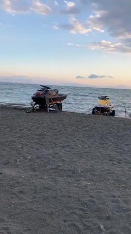 The video pans across a beach with dark sand. In the foreground, there are two jet skis parked on trailers. One jet ski is black and orange, and the other is yellow and white. Further down the beach, there is a low wooden table with two chairs, one of which is a green beanbag chair. The ocean is visible in the background with moderate waves. The sky is cloudy with hues of orange, pink, and blue, suggesting it is either sunrise or sunset. There are no people visible in the video.