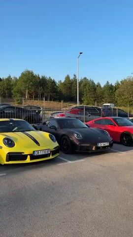 The video shows a parking lot filled with Porsches. The cars are parked in rows and are of various colors including yellow, dark brown, magenta, gray, and red. The environment appears to be outdoors during the day, with a clear blue sky and trees in the background. The cars are clean and in good condition. The video pans across the parking lot, showcasing the collection of Porsches. The license plates of the cars are visible in some shots, suggesting they are in a location with Bulgarian license plates. There are no people visible in the video.