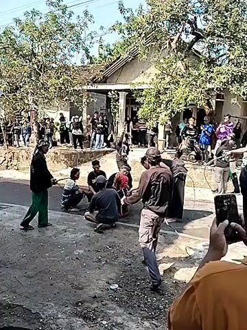 The video shows an outdoor scene with a group of people gathered on a street in front of a house. The atmosphere appears to be festive or ceremonial. Several individuals are engaged in what seems to be a performance or ritual involving whips. One person in the foreground is holding a whip and is in the process of cracking it. Others are seated or standing around, observing the activity. The environment includes trees, a building with a porch, and a paved or dirt road. The people are dressed in casual clothing, and some are wearing head coverings. There's a sense of community gathering, with spectators on the sides of the street.
