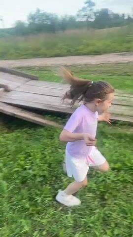 A young girl with dark hair in a ponytail, wearing a purple t-shirt and white shorts, runs across a grassy area. She is followed by a boy riding a dirt bike, who is wearing a white t-shirt and shorts. In the background, there are some buildings and trees. The sky is overcast.