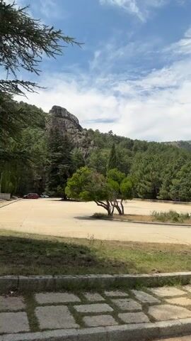 A vertical, medium-shot video pans left to right showing a paved area with grass and stone borders in the foreground. In the middle ground, a large, flat, light-colored paved area is visible with a small tree in the center. Behind this, a wooded hillside rises steeply with large rock formations at the crest. A tall, slender cross is visible on the left side of the frame, on the top of the hill. The sky above is blue with scattered white clouds. A red car is parked on the far left side of the paved area.