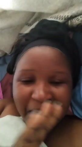 The video shows a woman lying in bed. She has dark hair pulled back with a black headband. She is smiling, looking into the camera, and saying "Hello guys, good morning." She is under the covers, and a small portion of her bare shoulder is visible. The bed has multiple layers of bedding with varied colors and patterns.