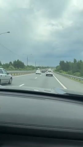 A first-person perspective shot from inside a car shows a highway with multiple lanes of traffic under a cloudy sky. The car is in the right-hand lane, following a white SUV. Several other cars, including a white sedan and a dark-colored SUV, are visible in front and to the left. Trees and green foliage line the sides of the highway, with a metal guardrail separating the road from the embankment. Streetlights are visible on the left side of the road. The dashboard of the car is in the foreground, obscuring the bottom portion of the view.