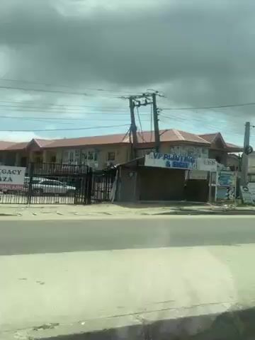 The video shows a view from inside a moving vehicle, driving along a paved road in a populated area. The sky is filled with dramatic, partly cloudy skies. Initially, the view includes a building with "VP PRINTING & SIGN" visible on its facade. As the vehicle moves, other buildings come into view, including residential houses with various roof colors and designs. A yellow and black auto rickshaw, also known as a "keke," passes in the opposite direction. At one point, the camera shifts slightly, bringing the person's hair into the foreground, partially obstructing the view of the outside scene. The focus then shifts back to the exterior, showing more of the street and buildings under the cloudy sky. Power lines are visible stretching across the sky.