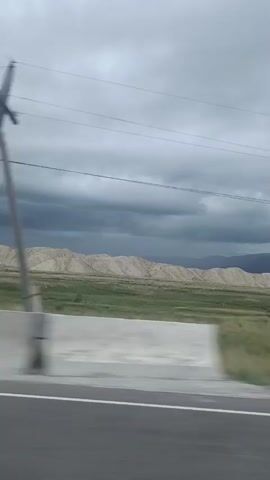 The video is a landscape shot from a moving vehicle on a cloudy day. It shows a grassy field with some scattered bushes and trees. In the background, there are rolling hills or mountains with a light-colored, sandy texture. A road with a white line is visible in the foreground, and the motion blur indicates the camera is in transit. Power lines and a utility pole are also present in the scene. The sky is overcast with dark clouds, suggesting impending rain or stormy weather. The overall impression is of a rural or remote, undeveloped area.