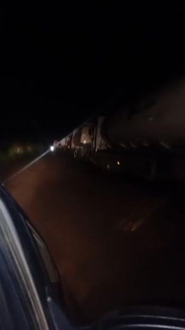 The video shows a long line of tanker trucks parked along a dark road at night. The camera is positioned inside a vehicle, looking out the side window at the trucks. The trucks are large and appear to be carrying some kind of liquid. There is a vehicle with its headlights on further up the road. The road surface appears to be paved but uneven. The overall environment is dark with limited lighting.