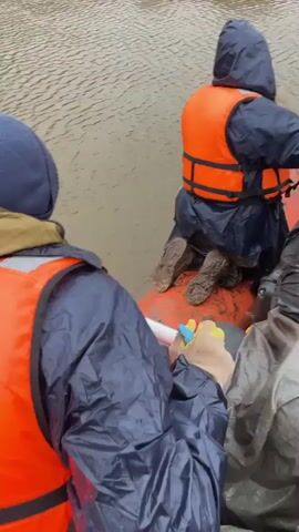 The video shows three or four people in a boat navigating a river on a cloudy day. The individuals are wearing life vests over their outer layers of clothing. One person wears a blue beanie, another an orange hood, and another a cap. The boat is red and gray. The surrounding environment consists of the river, with a visible bank lined with dry vegetation and bare trees. There are also tree branches that are partially submerged in the water. One of the individuals uses a paddle to navigate the boat along the water. The camera is positioned inside the boat looking out toward the river.