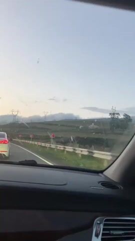 The video appears to be taken from the passenger seat of a car, looking out the windshield. The car is on a road, and there are other cars visible in front of it. The landscape outside is green and hilly, with power lines and utility poles visible in the distance. The sky is light blue with some clouds. The video appears to be taken during the day, likely in the late afternoon or early evening, as the light seems to be fading. There are no people visible other than the implied driver and passenger.