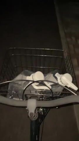 The video shows a first-person perspective of someone riding a bicycle at dusk. The bicycle has a wire basket on the front containing two plastic-wrapped items. The path ahead is dark, and the sky is a gradient of dark blue to orange. On the left is a multi-story building, and on the right is an overgrown area and a low-rise building with a light on. The bicycle is moving forward, and the camera is slightly shaky.