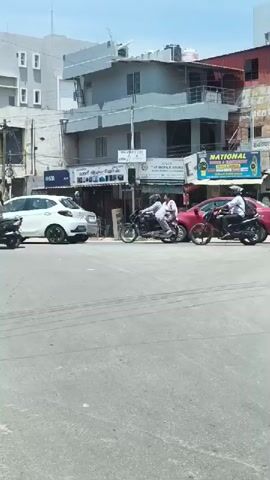 A street scene with several vehicles, including cars and motorcycles, on a sunny day. Two people on a motorcycle in the foreground are wearing helmets. A white car is parked to the left, and a red car is in the middle of the frame. Further down the street, a truck with a blue container is visible. The buildings in the background appear to be shops and residences.