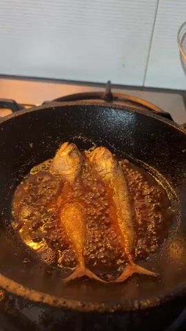The video shows two small fish frying in a black pan on a stovetop. The fish are golden brown and appear to be crispy. The oil in the pan is bubbling, indicating that the fish are cooking. The background is a kitchen with white tiles.