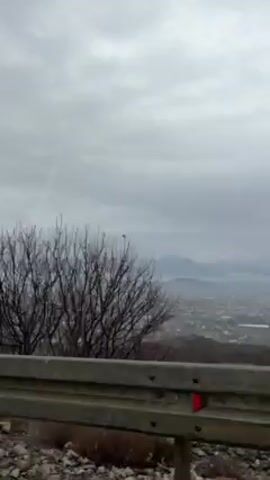 The camera faces a cloudy horizon over a valley full of houses, as seen from above a roadside guardrail. The valley is partially obscured by mist, and hills are faintly visible in the distance. A bit of bare brush can be seen beside the guardrail. The video is static.