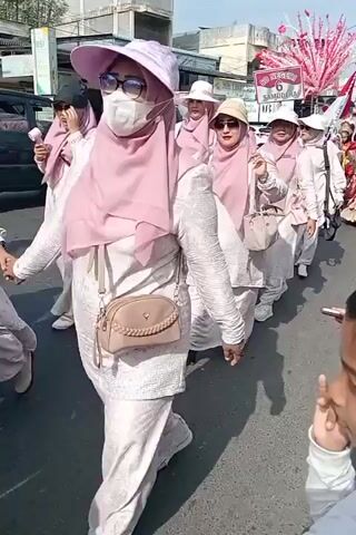 The video shows a parade or procession on a street. Several women wearing white and pink hijab and matching outfits are walking in the foreground and midground. They are also wearing hats and sunglasses. One woman in the midground is holding a light-colored purse. Behind them, a group of children in uniforms are walking. Some of the children are carrying flags, including an Indonesian flag, and a sign that says "SD NEGERI 6 SAMUDERA". There are decorative red and white structures on sticks being carried. Buildings line the street in the background. The sky is visible and appears to be sunny. The overall atmosphere seems festive.