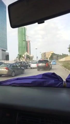 The video is shot from inside a car, looking forward through the windshield. The car appears to be in traffic, with other vehicles visible in front. A purple umbrella is draped over the dashboard in the foreground. In the background, there are several tall buildings, some of which appear to be under construction with cranes. The sky is visible and appears overcast. The overall scene suggests a busy urban environment.