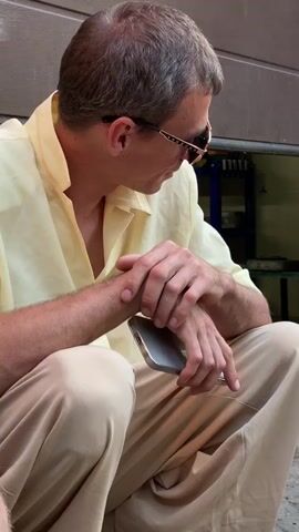 A man in a straw cowboy hat, a light yellow collared shirt, and burgundy pants is kneeling and using an orange orbital sander on a metal object. He is wearing white work gloves and dark sneakers. Another man, wearing a light yellow collared shirt and tan pants, is sitting and holding a silver smartphone in his lap. He is wearing sunglasses. In the background, there is a brick wall, a partially open garage door, and various tools, including a green drill and other sanders. The ground is uneven and appears to be concrete or dirt.