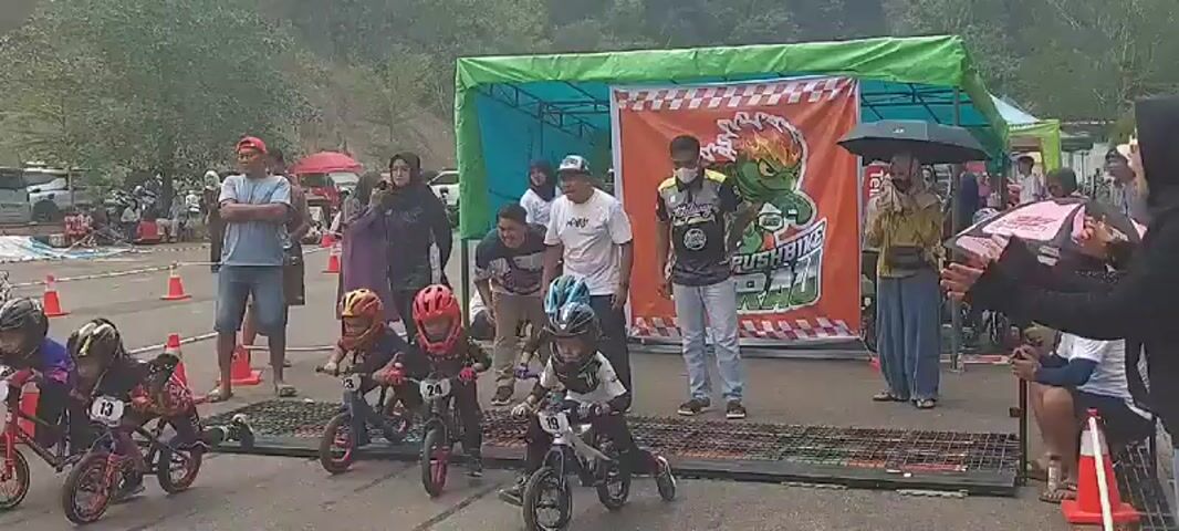 A children's balance bike race is underway on an asphalt track. Several young children, wearing helmets and protective gear, are riding balance bikes. Spectators, including adults and possibly older children, stand on the side of the track watching the race. Some spectators are holding cameras and phones, seemingly taking pictures or videos of the event. There are orange traffic cones and a checkered finish line tape. A banner is visible in the background with some text and an image of a cartoon character. The environment appears to be outdoors, with trees and a cloudy sky in the background.