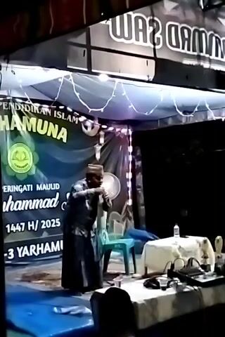 A man wearing a white brimless hat and a long, dark skirt-like garment stands on a stage and speaks into a microphone. He gestures with both hands. Behind him is a green banner, strung with white fairy lights, that says "PENDIDIKAN ISLAM HAMUNA."  Below that, the banner says "PERINGATI MAULID MUHAMMAD SAW 1447 H / 2025." Under the banner is a clear plastic chair and to the right of the man is a white table with a bottle of water and electronic equipment.  The stage is covered in a blue plastic tarp. Above the stage is a canopy with a banner that reads "RAMAD SAW." The event appears to be taking place outdoors at night.