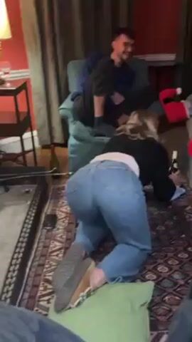 The video shows a woman on her hands and knees on a rug, reaching under a light-colored couch, possibly searching for something. She is wearing a black sweater, blue jeans, and slippers. A man is seated on the couch to the left, and another person stands to the right, recording with a phone. Two large red Santa boot decorations are visible on the couch next to the person filming. There is a half-empty bottle of wine on the floor near the couch, and other chairs are visible in the background. The room has red walls, curtains, and a fireplace.