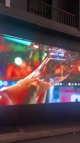 The video shows a night scene in Paceville, St Julian's, Malta. It begins with a close-up of a large outdoor LED screen advertising "The Long Hall Irish Pub." The screen displays an image of a champagne flute held by a hand, followed by the pub's logo and an invitation to visit, with an arrow indicating the direction. The camera then pans out to reveal the street scene. The street is paved and decorated with overhead string lights creating a festive atmosphere. Several people are walking around, some in groups. Buildings line the street, and signage for other establishments is visible. The overall impression is that of a lively nightlife area.