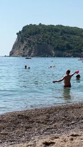 A man in red swim trunks stands in the shallow water near a pebble beach, facing a red ball that is in the air. He appears to be in the process of throwing or catching the ball. Further out in the ocean, two people are swimming, and another person is on a float. In the background, a forested cliff rises from the sea. The sky is clear and blue, indicating a sunny day.