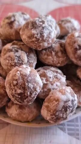 A close-up shot shows a pile of round, brown cookies dusted with white powder, likely powdered sugar. One cookie is being held up by a finger in the foreground, making it the focus of the image. The cookies are arranged on a plate that has a floral pattern. The background is blurred, but appears to be a table covered with a tablecloth.