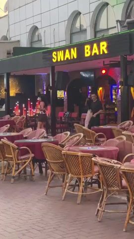 A man and a woman are dancing on the street in front of a bar called "SWAN BAR". The bar has outdoor seating with tables and chairs. The man is wearing a black shirt and white pants, and the woman is wearing a cow-print top and skirt. There are lights and speakers visible inside the bar. The street is paved with bricks.