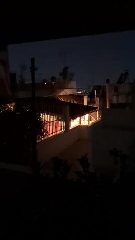 The video shows a dimly lit, outdoor scene at dusk or night. The view is from a slightly elevated position, looking out over rooftops. In the foreground, dark foliage is visible. The middle ground features several buildings with some illuminated windows and balconies, suggesting indoor activity. One building has a covered patio area with warm, artificial light. In the background, the night sky is visible, transitioning from a deep blue to black, with faint lights of a distant cityscape or other structures. The camera appears to be either stationary or panning slightly. The overall atmosphere is one of quiet observation of urban life at night.