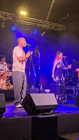 A band performs on a stage under a tent. A male singer in a light-colored t-shirt and grey pants stands at a microphone, gesturing with his hands. A female singer wearing a long, dark dress with a celestial pattern stands to his right, also at a microphone. Behind them, a drummer is partially visible. The stage is lit with blue and yellow lights, and there are speakers and other stage equipment visible.
