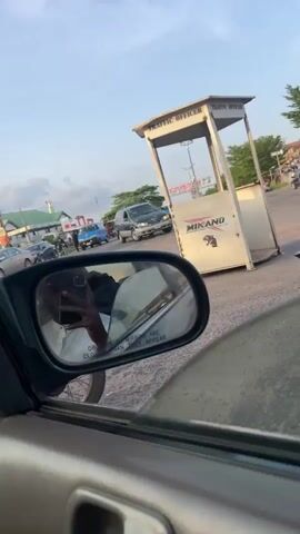 The video captures a dynamic street scene from the perspective of a vehicle, looking out the passenger side window and into the side mirror. The setting is Osogbo, Nigeria, during what appears to be late afternoon or early evening, evidenced by the warm, golden light and the low sun creating a glare towards the end of the clip.

The side mirror, prominently featured, reflects the person holding the camera (indicated by a hand gripping a phone) and parts of the car's interior. The common warning, "OBJECTS IN MIRROR ARE CLOSER THAN THEY APPEAR," is clearly legible on the mirror's surface.

Outside the car, a bustling urban environment unfolds. In the initial frames, several cars—including a silver SUV, a blue sedan, and a gray car—are visible on the road, suggesting moderate traffic or parked vehicles. A building labeled "TRAFFIC OFFICE" stands on the right. Further down the street, buildings with green roofs and what looks like a mosque minaret can be seen against a partly cloudy sky.

As the camera pans to the right, more of the street life comes into view. Buildings adorned with red and white banners, possibly advertisements, line the road. People are seen walking on the sidewalks, and market stalls covered with tarpaulins are set up, indicating commercial activity. One man in a dark tunic is seen walking past the car. The scene culminates with the bright, low sun casting a strong glare into the frame, illuminating more distant market activities and pedestrians.

The overall atmosphere is lively and typical of a busy street in a Nigerian city, with a mix of vehicle movement, pedestrian traffic, and roadside commerce under the soft glow of the setting sun.