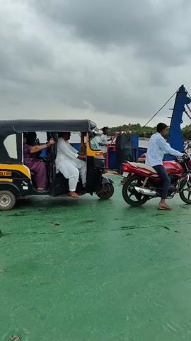 A video shows a ferry with several vehicles on its deck. On the left, a three-wheeled auto-rickshaw carries four passengers. In the center, a person on a red motorcycle faces away from the camera. To the right, three people walk towards the back of the ferry. Further back, several motorcycles are visible, and the ferry appears to be approaching a shore with vegetation and buildings. The sky is overcast.