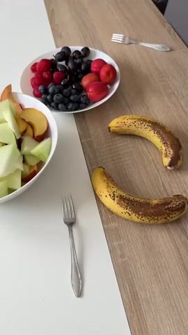 The video pans across a wooden table covered with a white runner. On the left side of the runner, a white bowl contains chopped melon and peaches. To the right of the bowl, a silver fork rests on the runner. Further to the right, a white plate holds a colorful assortment of fruits, including blueberries, raspberries, grapes, and small red fruits. Two ripe bananas lie on the wooden surface to the right of the plate. The background shows a blurred living room area with furniture and decor.
