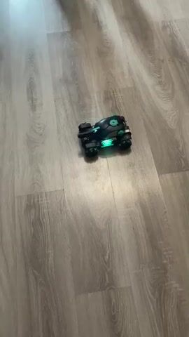 A toy remote-control car, black with green lights, moves across a light wood-grain floor. The car is positioned in the center of the frame. In the upper left, a portion of a wooden shelf or toy is visible. A person's dark shoe is seen at the top right of the frame in the last two seconds. The car appears to be moving forward and slightly to the right.