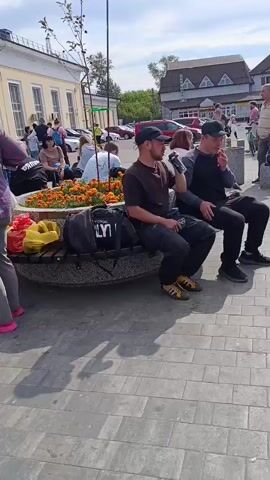 The video shows a sunny outdoor scene with several people and buildings. In the foreground, a person in a pink plaid shirt and gray pants is standing, with bright pink shoes. To their right, two men are sitting on a bench. One man in a dark shirt and cap is holding a phone to his ear, while the other man in a dark jacket with blue stripes on the sleeve is sitting next to him. A large black bag and a yellow bag are on the bench. Behind them is a flower bed with orange flowers and a small tree. In the background, there are buildings with yellow facades and signs in Russian. One sign reads "КОФЕЙНЯ" (cafe), and another reads "ТАБАК" (tobacco). Further down, there is a shop with the sign "СКУПКА" (pawnshop) and "ПОКУПКА ПРОДАЖА ОБМЕН" (buy sell exchange). A pink scooter with a basket is on the ground in the middle of the paved area. People are walking around, some with luggage.