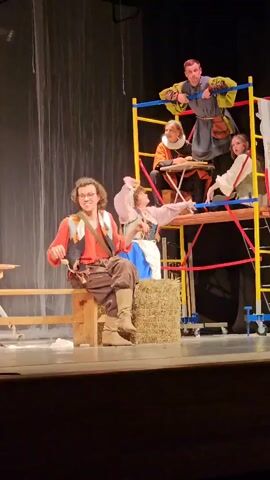 A group of actors is on a stage, possibly performing a play. In the foreground, a man with curly brown hair and glasses sits on a wooden stool. He wears a reddish-orange shirt, a dark vest with decorative trim, and loose brown pants tucked into light brown boots. He holds a small object, possibly a prop, in his right hand. A light brown bag is slung across his body. To his right, a bale of hay sits on the stage.

Behind the man and the hay bale, a scaffolding structure is set up. On a lower platform of the scaffolding, a woman in a pink and white outfit with a blue skirt is seated. She gestures with her right hand. On a higher platform of the scaffolding, three other actors are visible. One man in a yellow and blue costume stands, looking down. Another person, also in a colorful costume, is seated at a small table, possibly playing a game or interacting with someone off-camera. A third person, wearing a white garment, is also on this platform.

The backdrop consists of a light-colored curtain. The lighting suggests a stage environment, with the main action illuminated and the background somewhat dimmer. The stage floor is visible in the foreground.