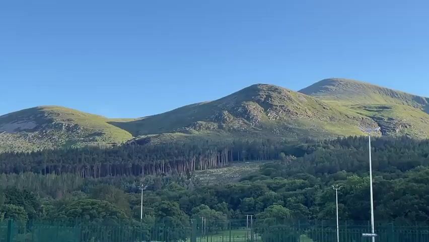 The video shows a wide shot of rolling green mountains under a clear blue sky. In the foreground, there is a fence and some green grass. In the middle ground, there is a dense forest of dark green trees, with some lighter green trees scattered throughout. There are also some power lines and poles visible in the distance. The mountains are covered in grass and rocks, and appear to be quite large. The overall scene is peaceful and scenic.
