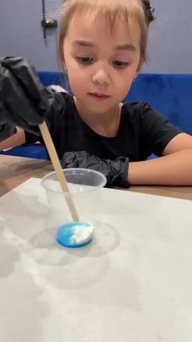 This media file features a close-up shot of a young girl, likely of Russian ethnicity given the location context of Murom, Russia, engaged in an art or craft activity. She is wearing a black t-shirt and black gloves, suggesting she is working with materials that require protection. The girl is focused on a clear plastic cup containing a mixture of blue liquid and white powder, which she is stirring with a wooden stick. Her expression conveys concentration. The background is slightly blurred, showing hints of a room with a wooden table and a blue upholstered seat. The lighting suggests it is daytime, possibly indoors in a well-lit space. The overall scene depicts a moment of creative engagement and learning for a child. There is no visible text within the media file.