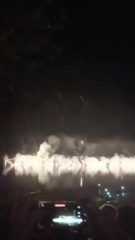 The video captures a nighttime fireworks display over a body of water. In the foreground, silhouetted figures, likely people, are visible, with one person holding a smartphone to record the event. The fireworks are exploding in the distance, creating bright bursts of light and plumes of smoke that are reflected in the water. The overall scene is dark, illuminated primarily by the light of the fireworks.