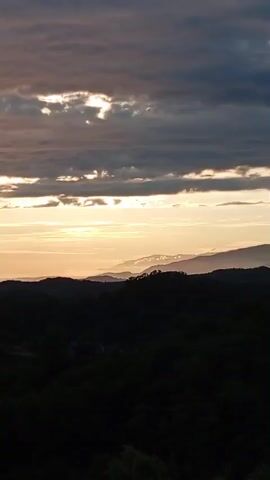 A time-lapse video captures a dusky mountain landscape. Silhouetted trees and hills fill the foreground, transitioning into layered mountain ranges in the mid-ground. A band of soft, golden light stretches across the horizon, indicating sunset or sunrise. Dark, textured clouds dominate the sky, with hints of brighter light peeking through them. The overall impression is of a serene, natural environment under changing atmospheric conditions.