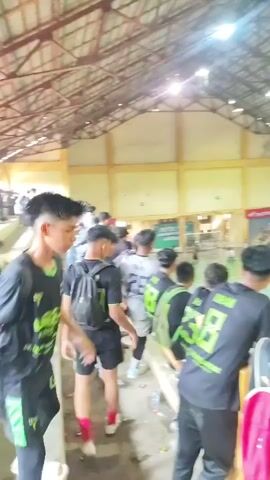 The video shows an indoor sports arena, likely a gymnasium, with a badminton court in the center. Spectators are seated in bleachers on two sides of the court, with the camera positioned among them. The spectators in the foreground are predominantly young males, many wearing black shirts with green accents. Some have backpacks. On the court, athletes, also dressed in sports attire, are engaged in a game, possibly badminton, given the court markings and net. Banners and advertisements are visible on the walls of the arena. The lighting appears to be artificial, coming from overhead fixtures. The atmosphere suggests a sporting event with an active audience.